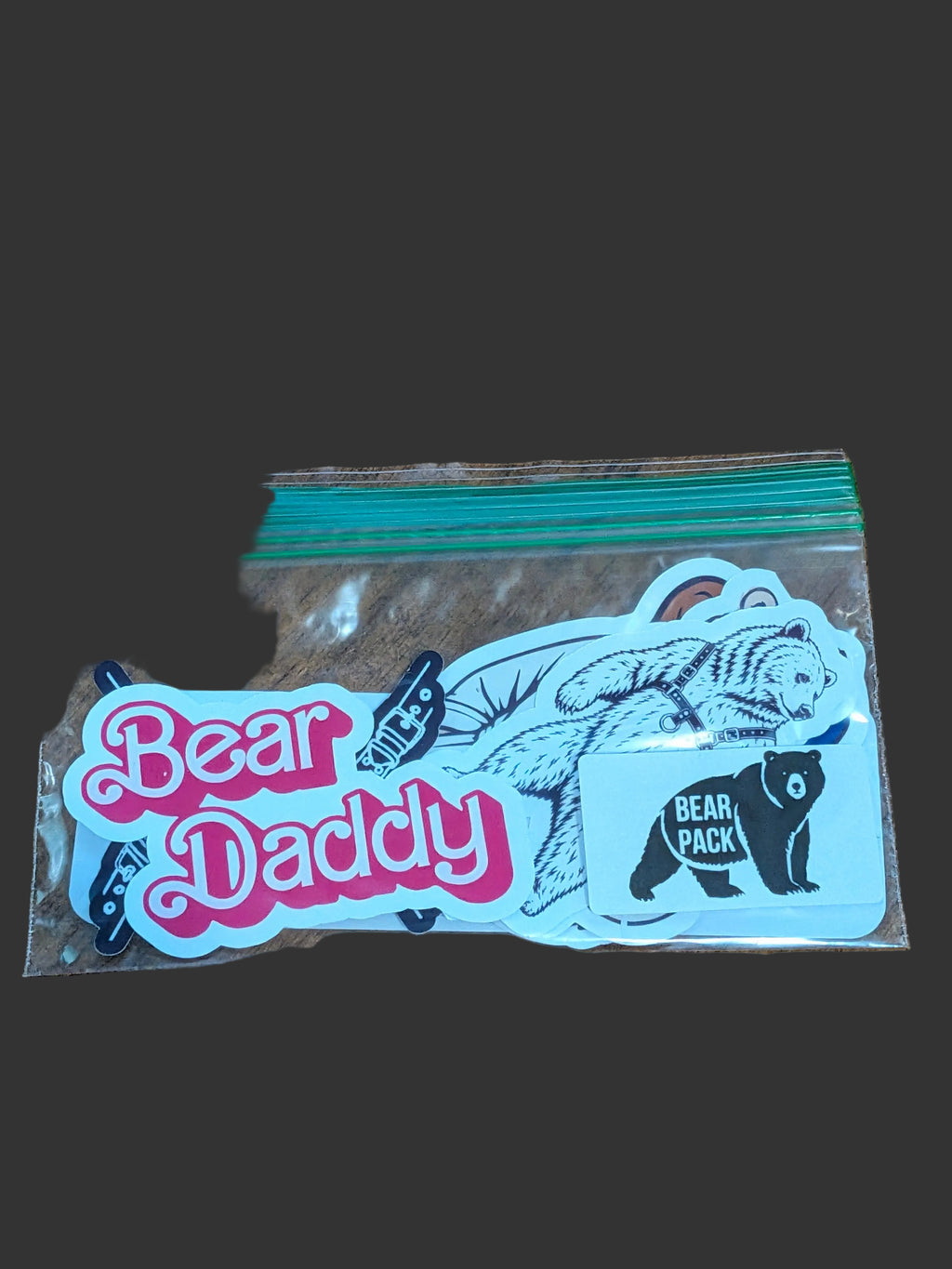 A clear plastic zip-top bag containing several die-cut stickers on a brown wooden surface. The stickers include a large pink and white 'Bear Daddy' script sticker, a black bear illustration with a blue bow, and a small black bear sticker labeled 'BEAR PACK'.